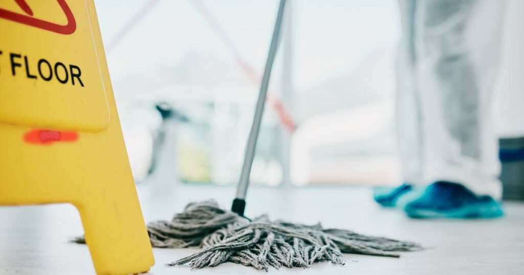 A person stands beside a wet mop and a sign reading "Floor Cleaning," illustrating the concept of using commercial cleaners.